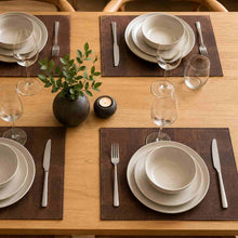 Load image into Gallery viewer, A styled dining table setting with four place settings, including stacked white ceramic plates, silver flatware, clear wine glasses, and dark brown cork placemats. A small dark textured vase with green leaves and a black candle sit in the center on a light wood table.